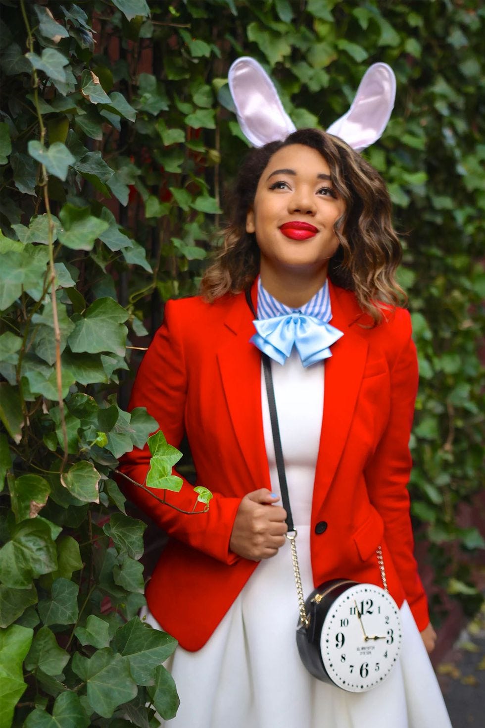 woman dressed up as the white rabbit from alice in wonderland standing against a wall covered with fig leaves and vines