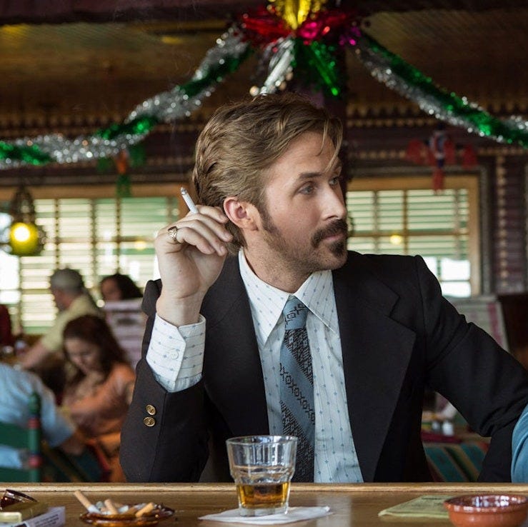 ryan gosling sits at a festive bar wearing a suit in a scene from the nice guys