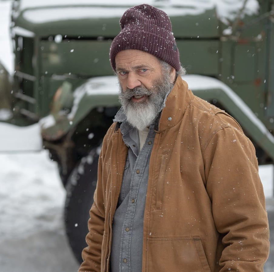 a man in a hat and coat stands in front of a military vehicle in a scene from fatman