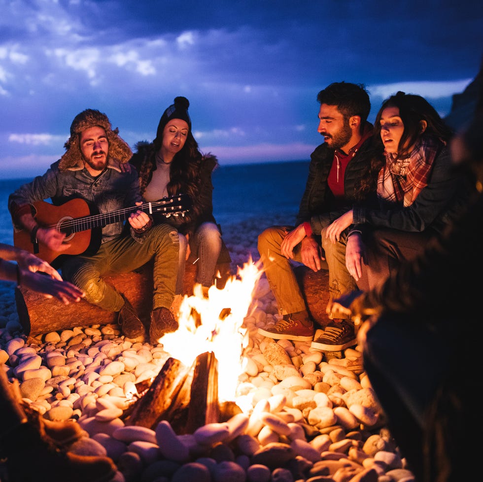 young hipster friends with a guitar singing around a campfire at a beach at dusk